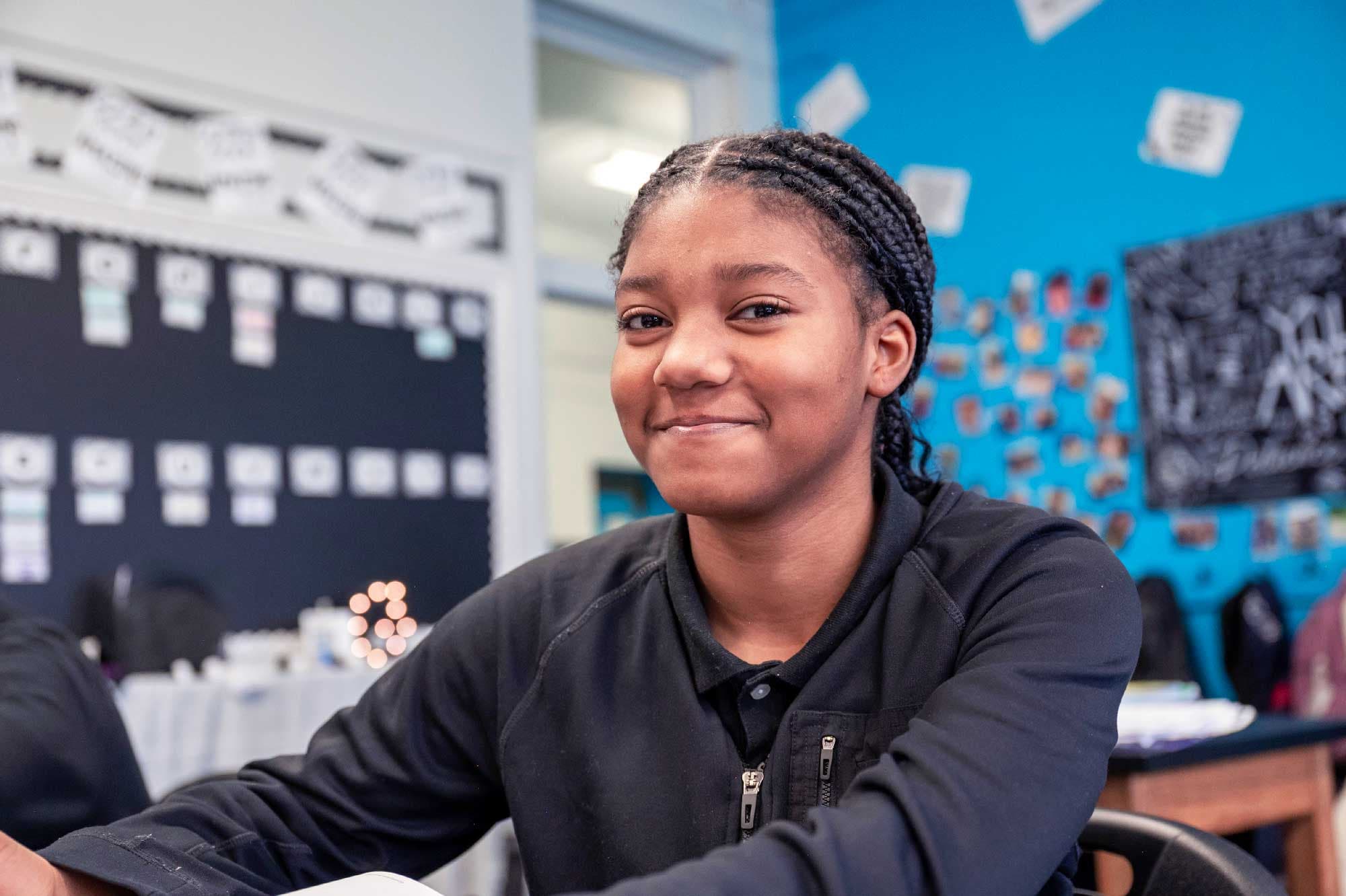 Smiling student with braided hair seated at a classroom desk.