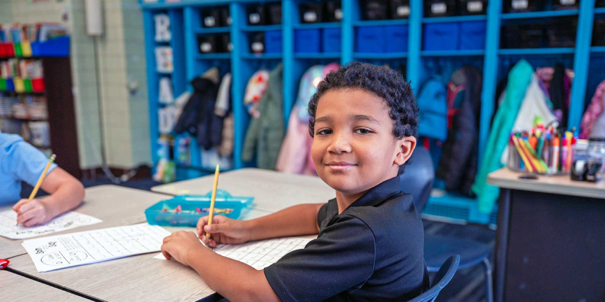 Student smiling while writing on a worksheet at a classroom desk.